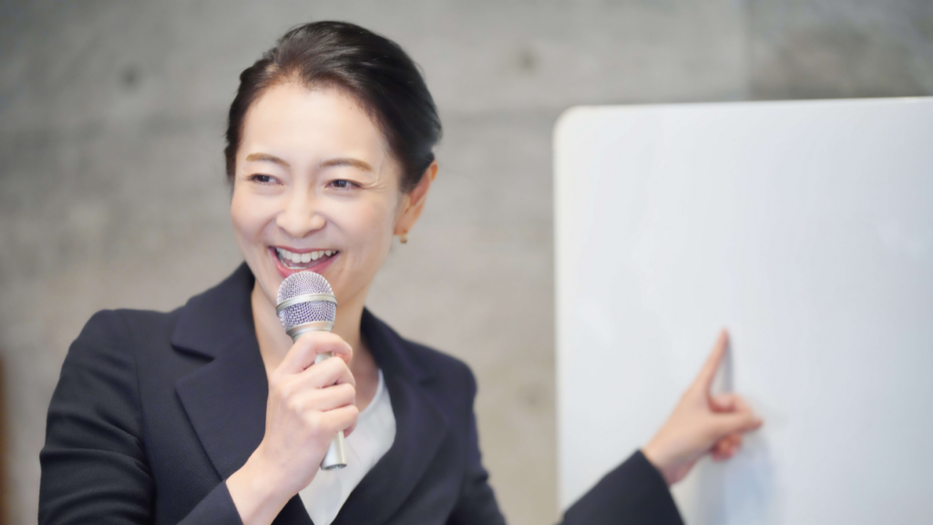 ホワイトボードを指差しながらマイクで話す女性